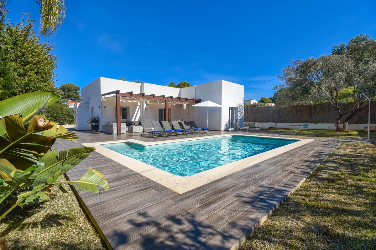 Villa blanca de diseño moderno con piscina rectangular, terraza de gres porcelánico efecto madera y porche con pérgola y mobiliario exterior.