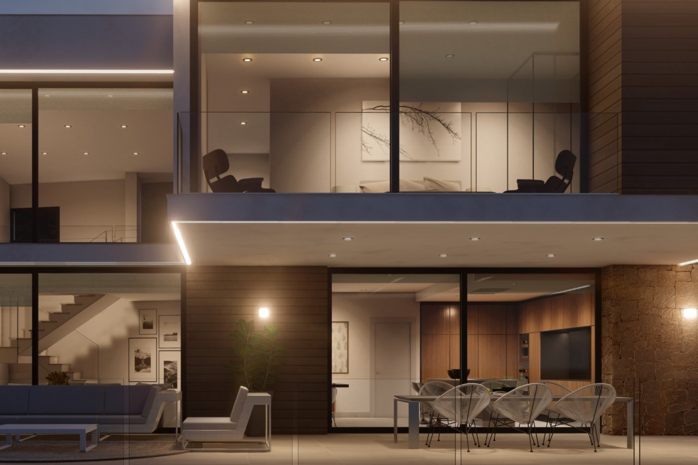 Vista exterior nocturna de una villa con terraza, sillas Acapulco y muros de piedra. Los ventanales muestran la cocina integrada y un balcón superior con barandilla de vidrio.