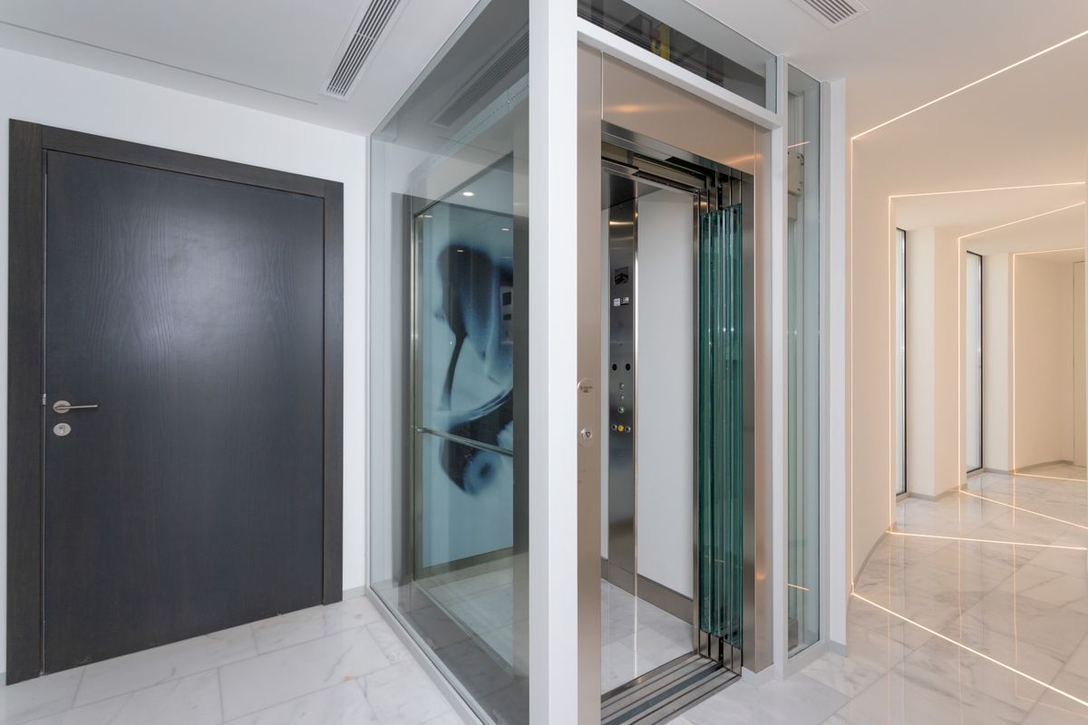 Ascensor moderno de cristal y acero inoxidable, situado sobre suelos de mármol blanco junto a un pasillo con iluminación LED lineal integrada.