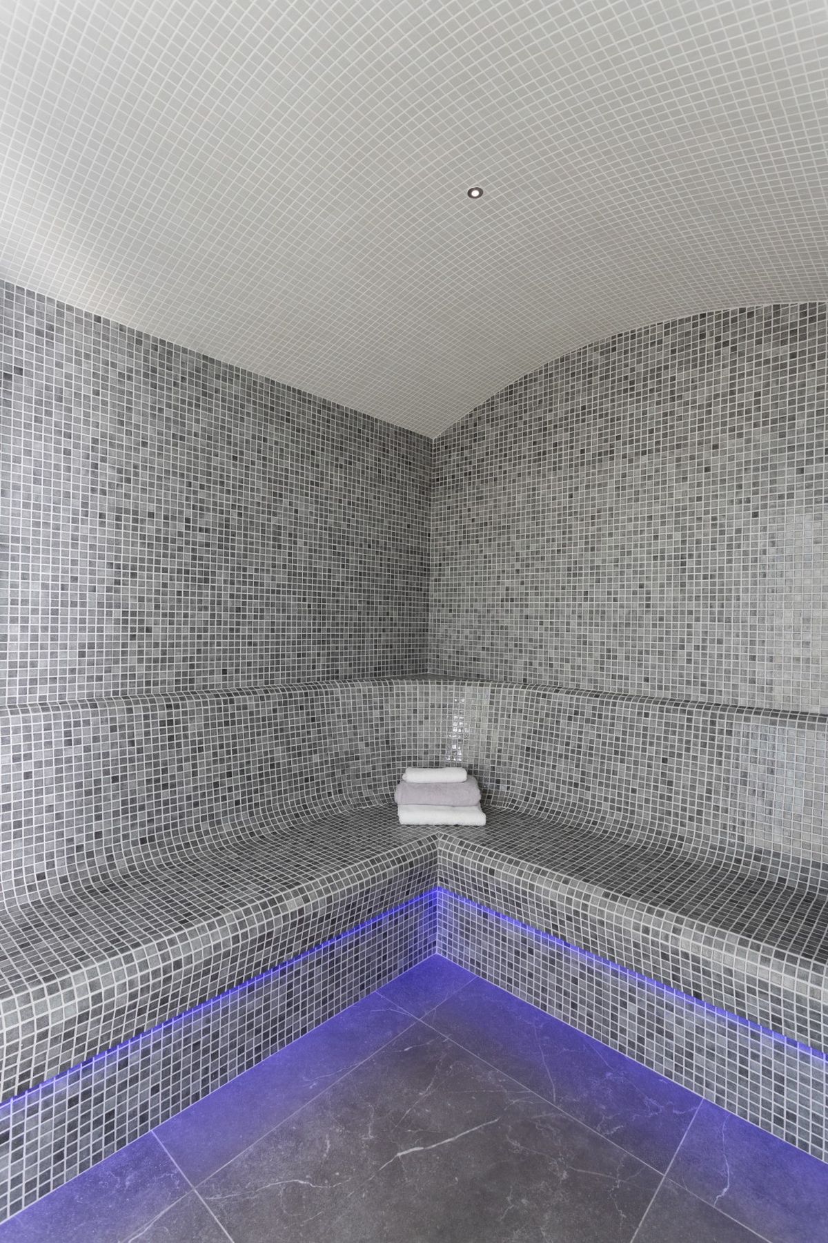 Baño de vapor con revestimiento de mosaico gris, bancos ergonómicos con iluminación LED azul y techo abovedado de teselas blancas.