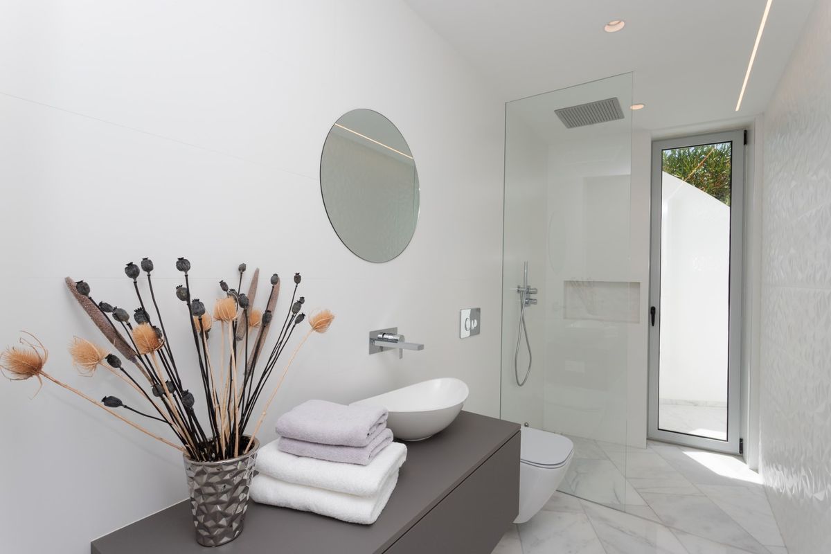 Baño moderno con lavabo sobre encimera gris, suelo de mármol blanco y ducha de obra con mampara de cristal y acceso directo al exterior.