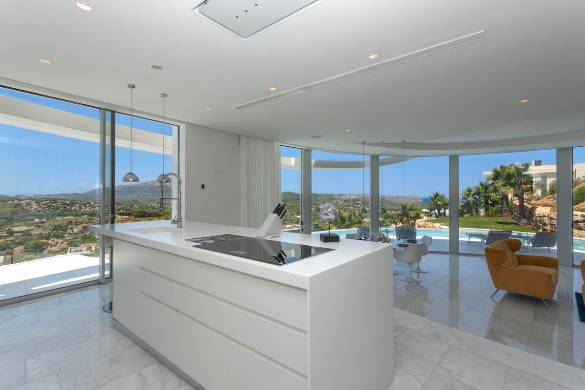 Cocina moderna de planta abierta con isla blanca, suelos de mármol y ventanales de suelo a techo que ofrecen vistas panorámicas al mar y la piscina.