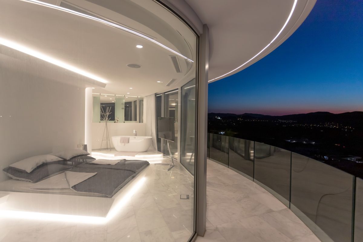Dormitorio minimalista con suelos de mármol, iluminación LED integrada, bañera exenta y terraza curva con vistas panorámicas al valle durante el anochecer.