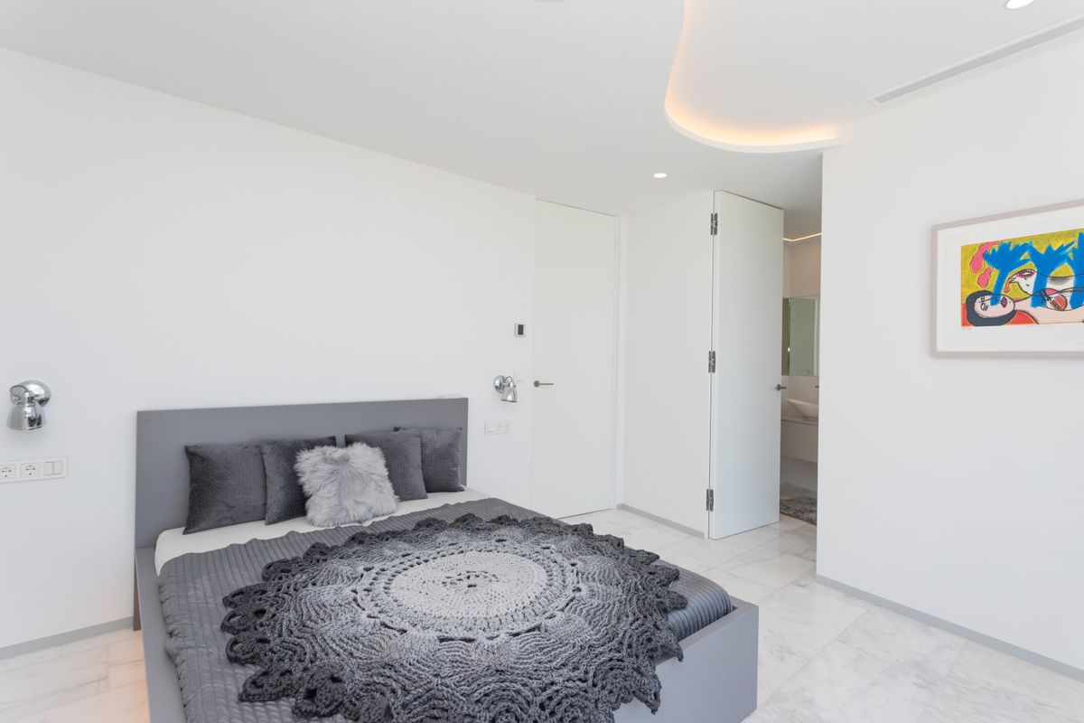 Dormitorio moderno con cama gris, suelos de mármol y techo con iluminación indirecta curva. Se aprecia el acceso al baño privado y una obra de arte contemporáneo.