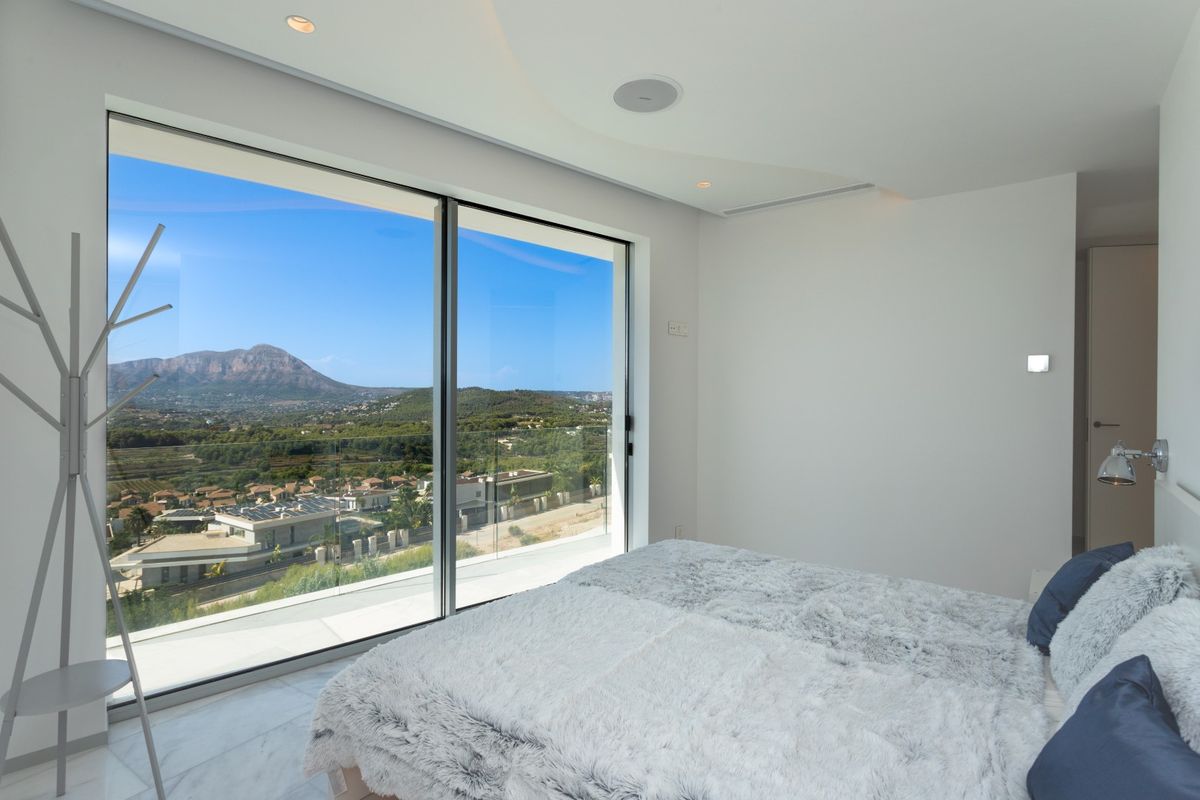 Dormitorio moderno con ventanales de suelo a techo y vistas al Montgó. Cuenta con suelos de mármol, iluminación empotrada y altavoces integrados en el techo.