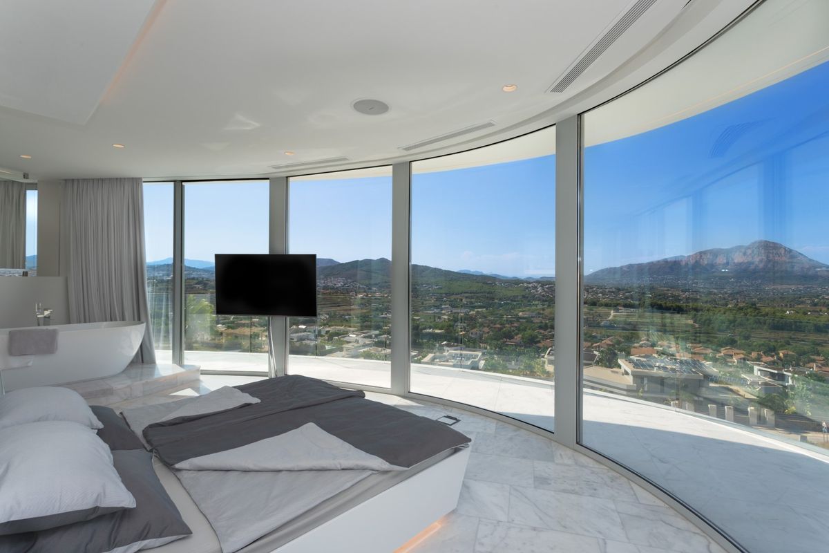 Dormitorio principal con ventanales curvos de suelo a techo, vistas panorámicas al valle, suelo de mármol blanco y bañera exenta integrada.