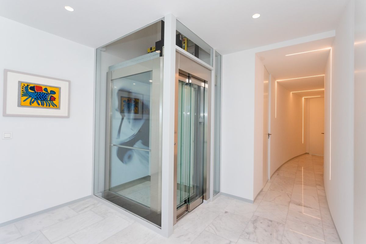 Pasillo moderno con ascensor acristalado y estructura de acero, suelos de mármol y un corredor iluminado mediante líneas de LED integradas en el techo.
