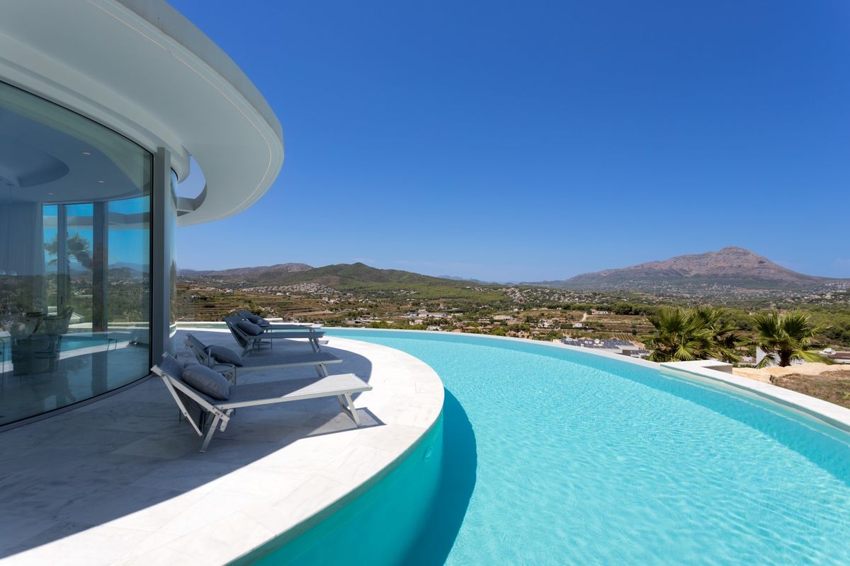Piscina infinita curva con agua turquesa y terraza de piedra blanca con tumbonas grises, ofreciendo vistas panorámicas al valle y las montañas.