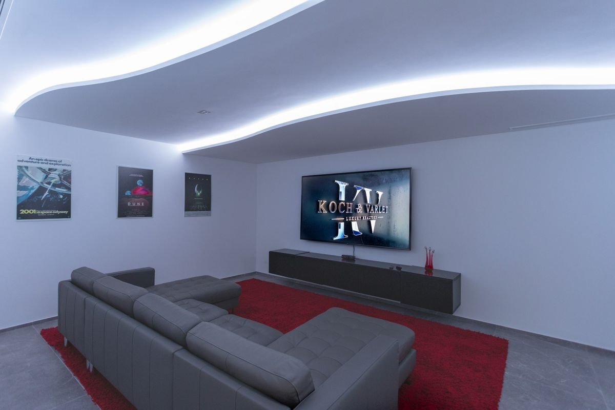Sala de cine privada con techo de diseño ondulado e iluminación LED indirecta, equipada con un sofá de cuero gris, alfombra roja y pantalla de gran formato.