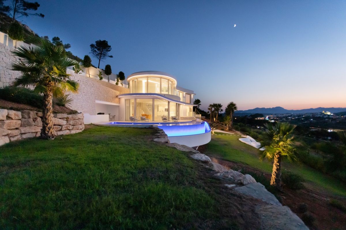 Villa moderna de diseño circular al anochecer con fachadas de vidrio curvas, piscina infinita iluminada y jardines aterrazados de piedra con vistas al valle.
