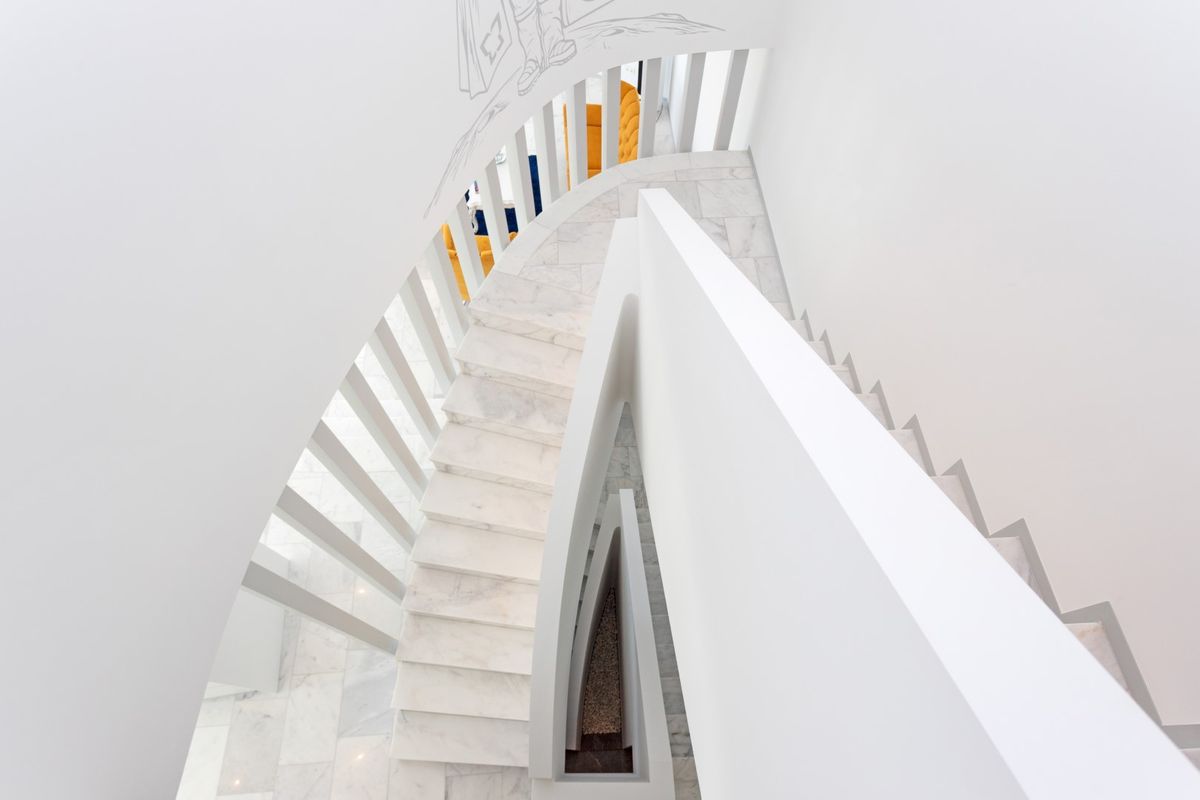 Vista cenital de una escalera curva minimalista con peldaños de mármol blanco y una balaustrada de listones verticales integrados en paredes blancas.