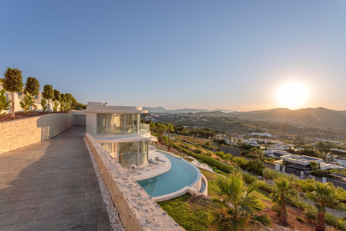 Vista exterior de una villa moderna con piscina infinita curva, fachadas de cristal y muros de piedra con vistas al valle durante el atardecer.