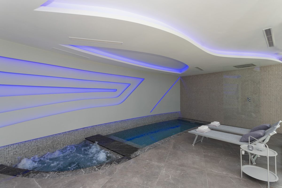Zona de spa interior con piscina de hidromasaje, iluminación LED perimetral en tonos azules, suelos de piedra gris y tumbonas de diseño contemporáneo.