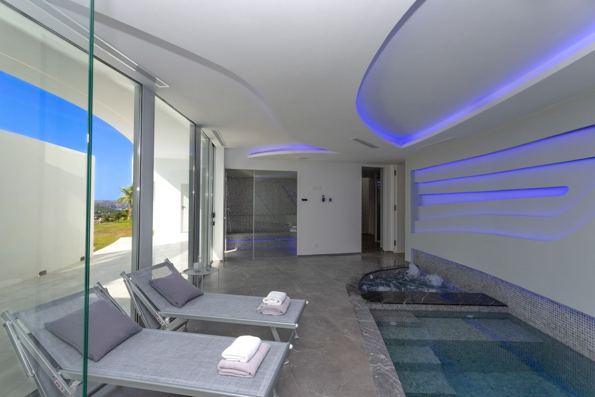 Zona de spa interior con piscina de mosaico, jacuzzi integrado y baño de vapor. Dispone de tumbonas grises, iluminación LED azul y amplios ventanales con vistas al exterior.