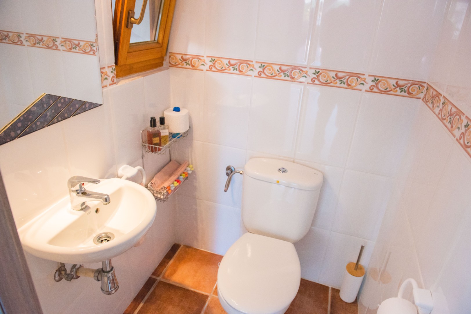 Aseo compacto con azulejos blancos y cenefa decorativa, suelo de terracota, lavabo cerámico de pared y ventana con marco de madera para ventilación natural.