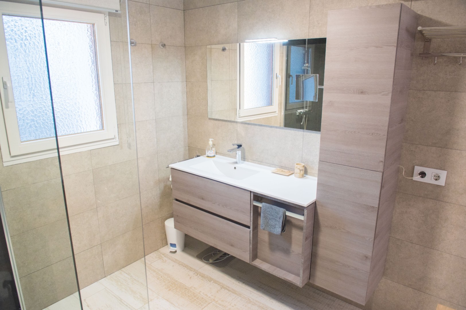 Baño moderno con mueble suspendido en acabado madera, lavabo integrado, columna de almacenaje a juego y paredes revestidas con azulejos en tonos arena.