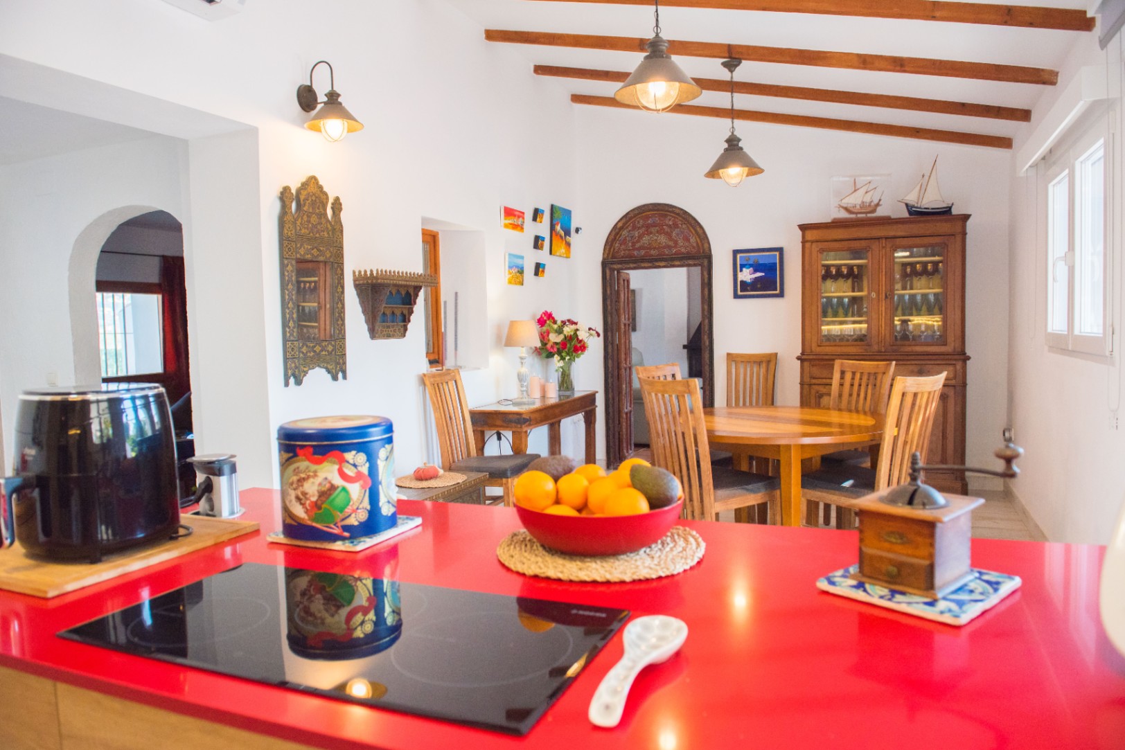 Cocina abierta con encimera roja vibrante y placa de inducción, integrada con un comedor rústico que destaca por sus vigas de madera vistas y mobiliario clásico.