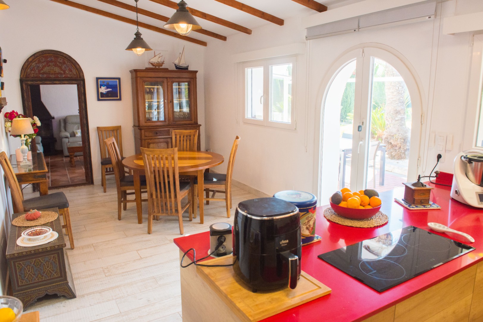 Cocina con isla roja y comedor integrado, vigas de madera a la vista y acceso directo al jardín mediante puerta de arco acristalada.