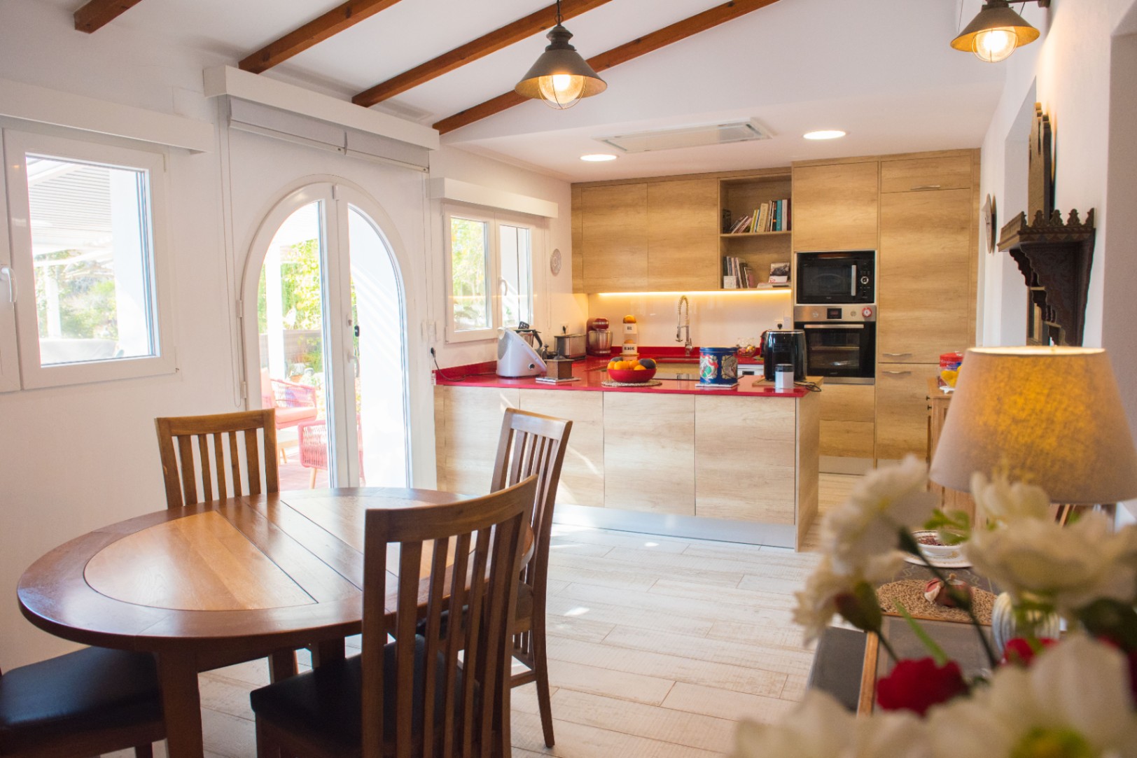 Cocina moderna con armarios de madera clara, encimera roja vibrante y electrodomésticos integrados, destacando techos inclinados con vigas de madera y zona de comedor.