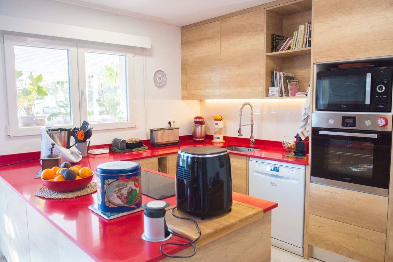 Cocina moderna con encimera roja vibrante, armarios de madera clara y electrodomésticos integrados, incluyendo horno, microondas y lavavajillas.