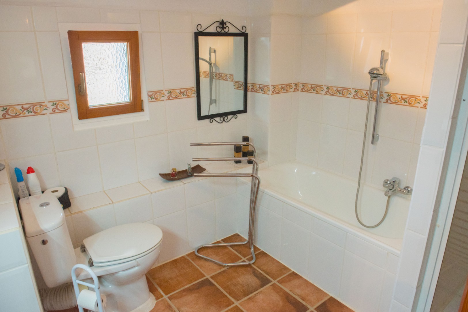 Cuarto de baño con bañera integrada, suelos de terracota y azulejos blancos con cenefa decorativa. Incluye una pequeña ventana de madera y espejo de forja.