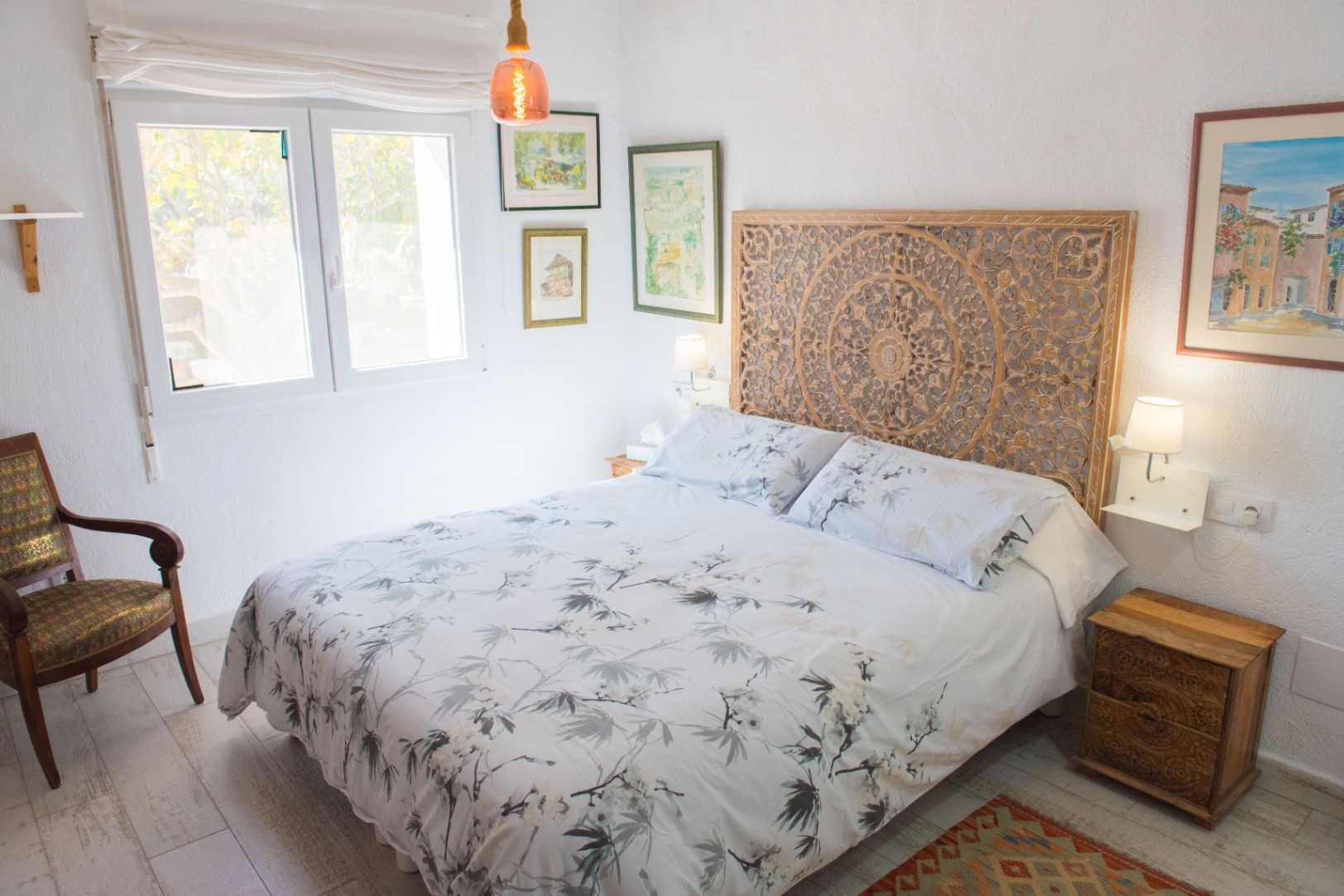 Dormitorio con cabecero de madera tallada, ropa de cama con motivos naturales y sillón vintage. Incluye suelos de gres imitación madera y cuadros decorativos.