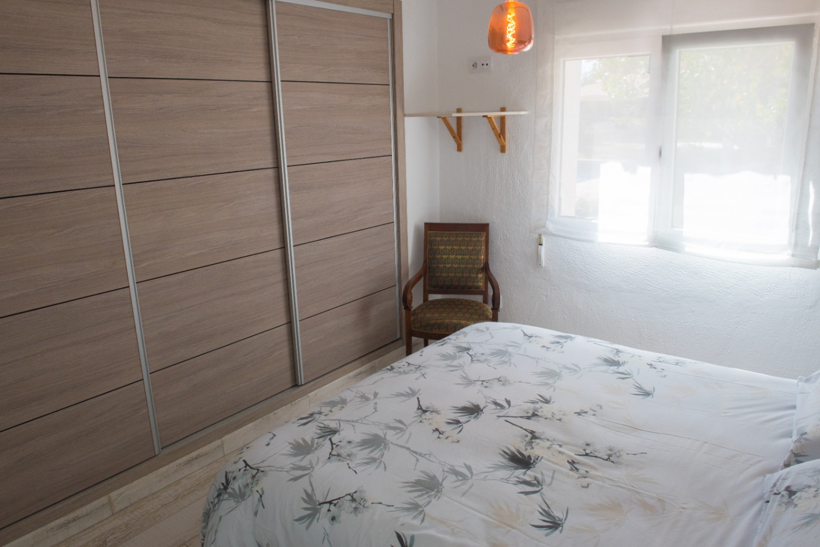 Dormitorio con un amplio armario empotrado de puertas correderas en acabado madera, un sillón clásico y luz natural filtrada por cortinas blancas.