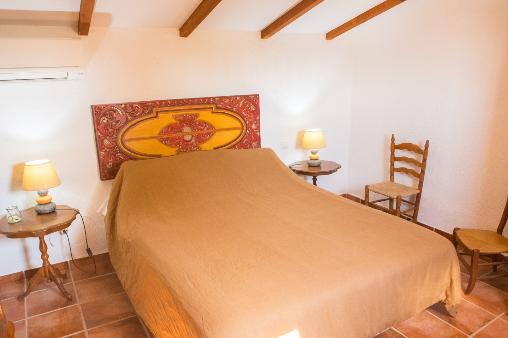 Dormitorio con un cabecero de madera tallada artesanal, vigas de madera a la vista, suelos de terracota y mobiliario rústico tradicional.