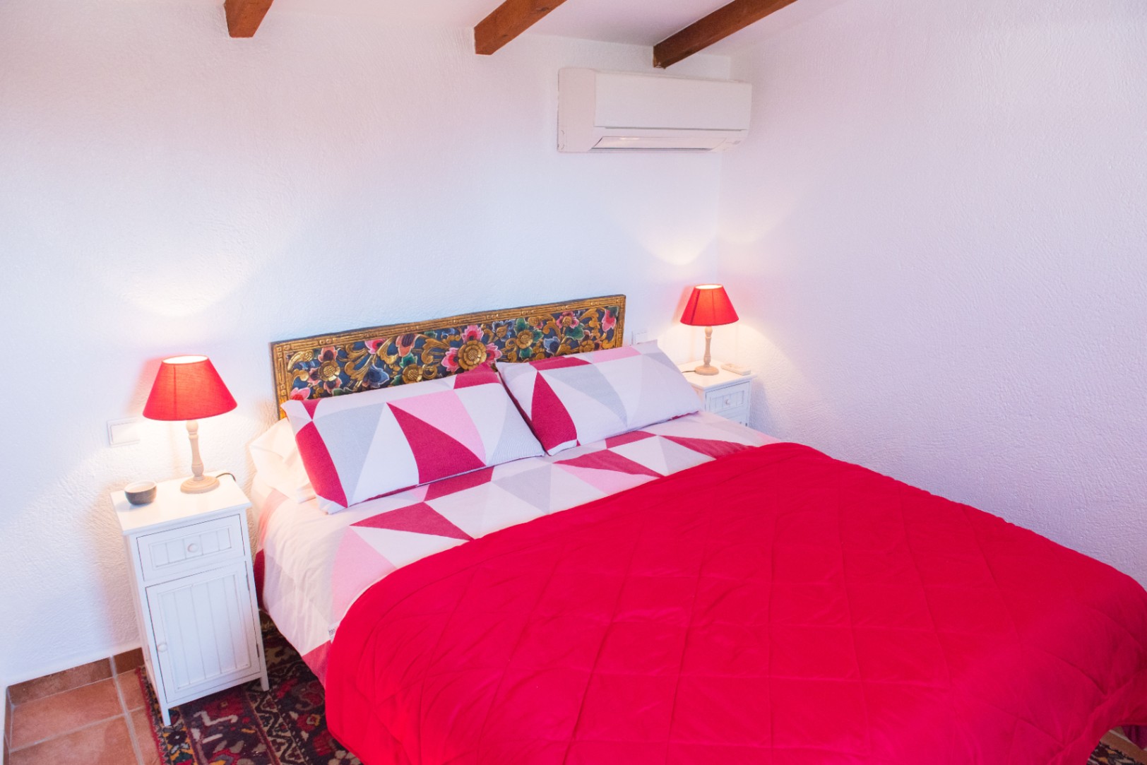 Dormitorio con vigas de madera a la vista, cabecero tallado artesanal y mesitas blancas con lámparas rojas. Incluye aire acondicionado y suelos de terracota.
