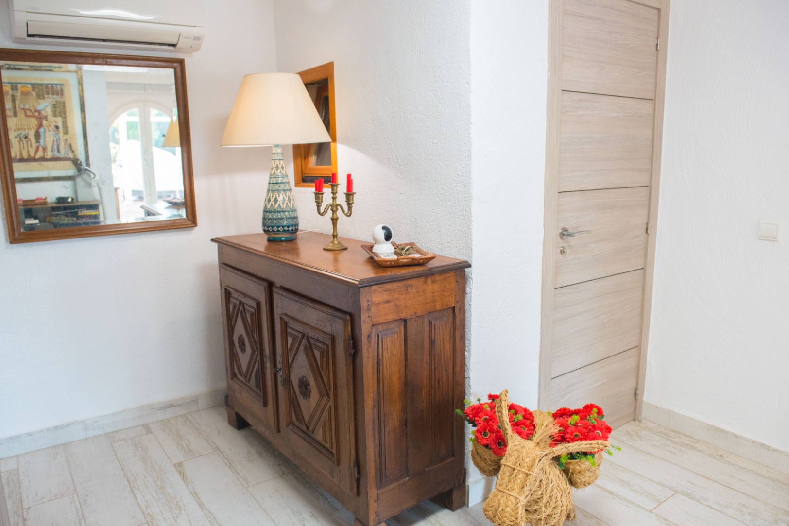 Entrada con aparador de madera tallada, lámpara de cerámica y suelos cerámicos de imitación madera. Incluye un espejo enmarcado y aire acondicionado sobre paredes blancas.