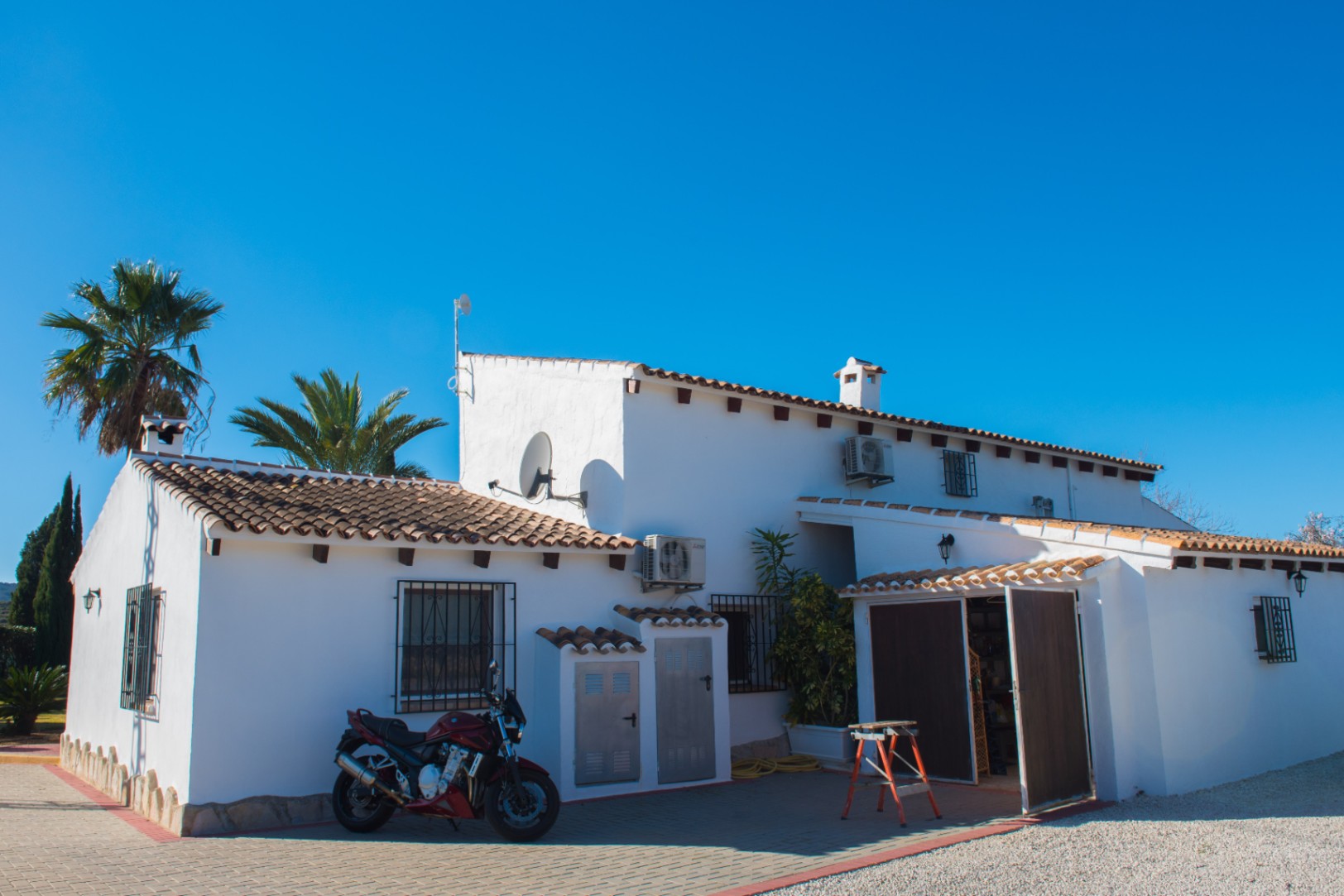 Fachada exterior de una villa blanca con tejado de teja cerámica, vigas de madera a la vista y patio pavimentado bajo un cielo despejado.