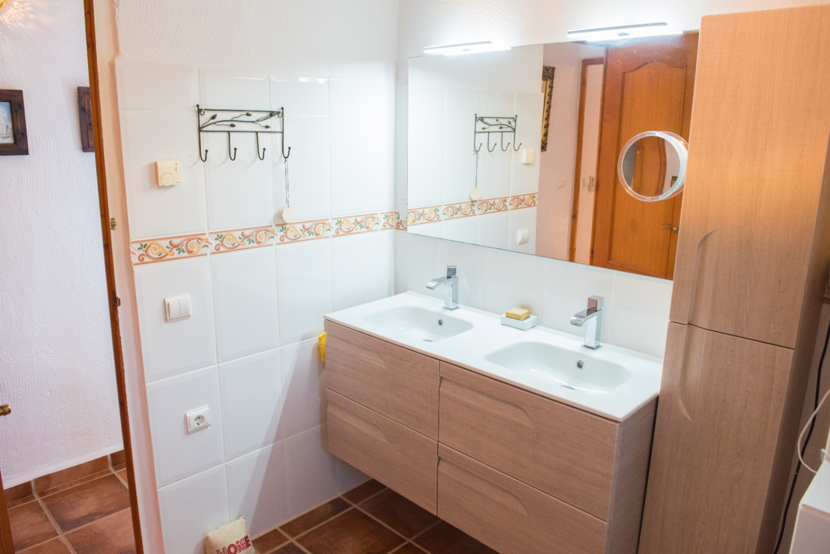 Mueble de baño suspendido en madera clara con lavabo doble integrado, grifería cromada y un amplio espejo con iluminación superior y espejo de aumento.