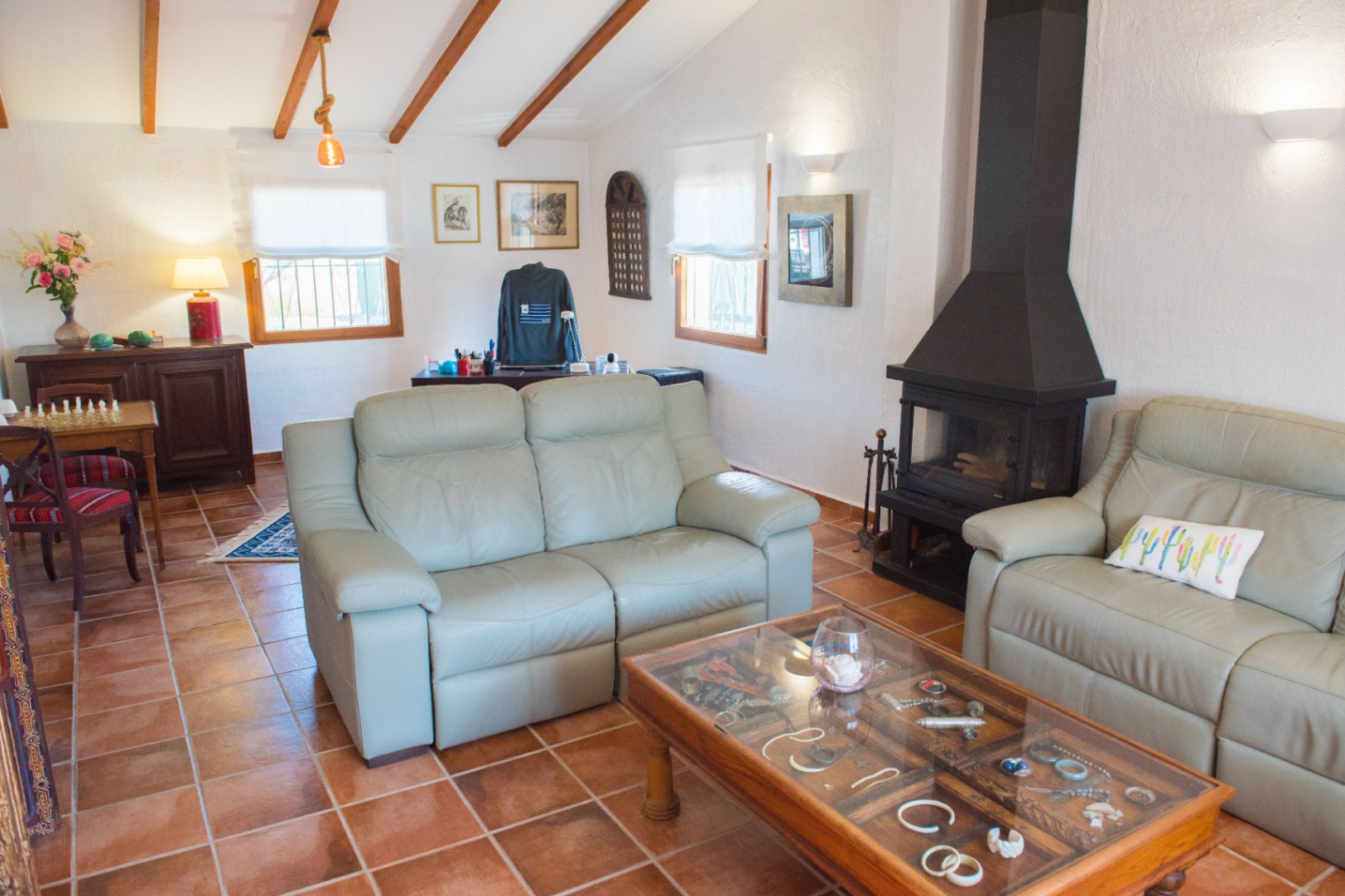 Salón con suelo de baldosas de terracota, chimenea de metal negro y techos inclinados con vigas de madera vistas. Dispone de sofás de cuero y una mesa de centro expositora.