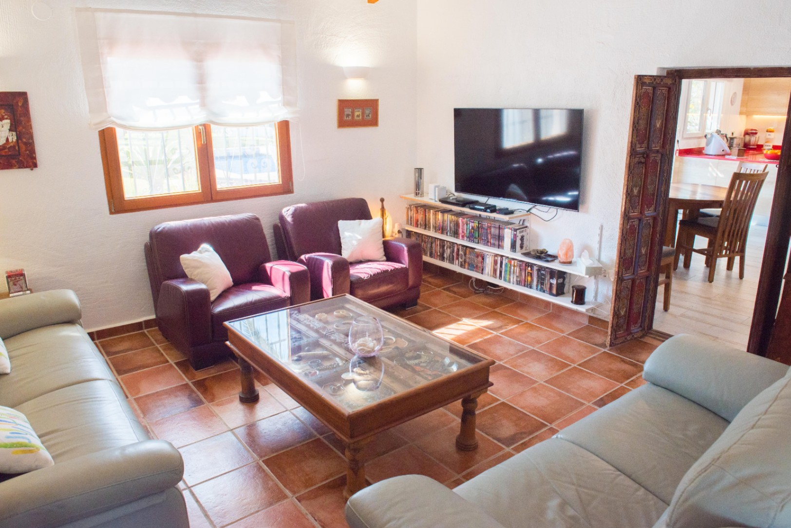 Salón con suelos de terracota, sofás de cuero y mesa de centro de cristal. Dispone de estantería multimedia bajo el televisor y acceso decorado hacia el comedor.