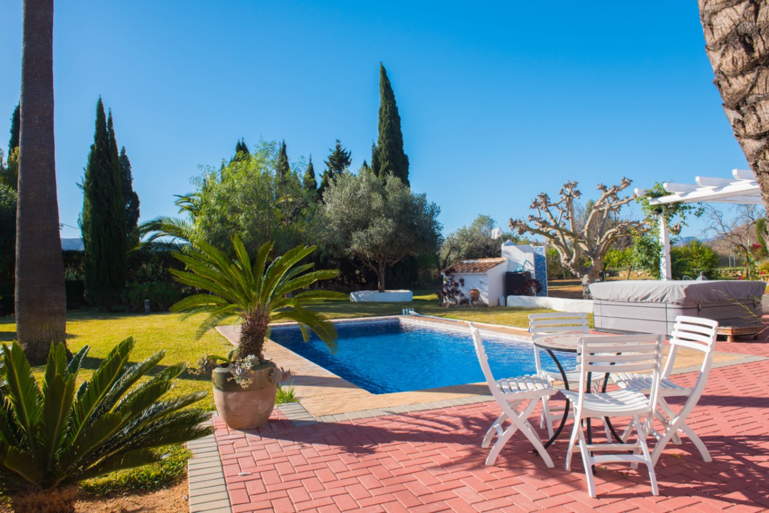 Terraza exterior con pavimento de ladrillo rojo, piscina rectangular, mobiliario de jardín blanco y zona ajardinada con cipreses y palmeras.