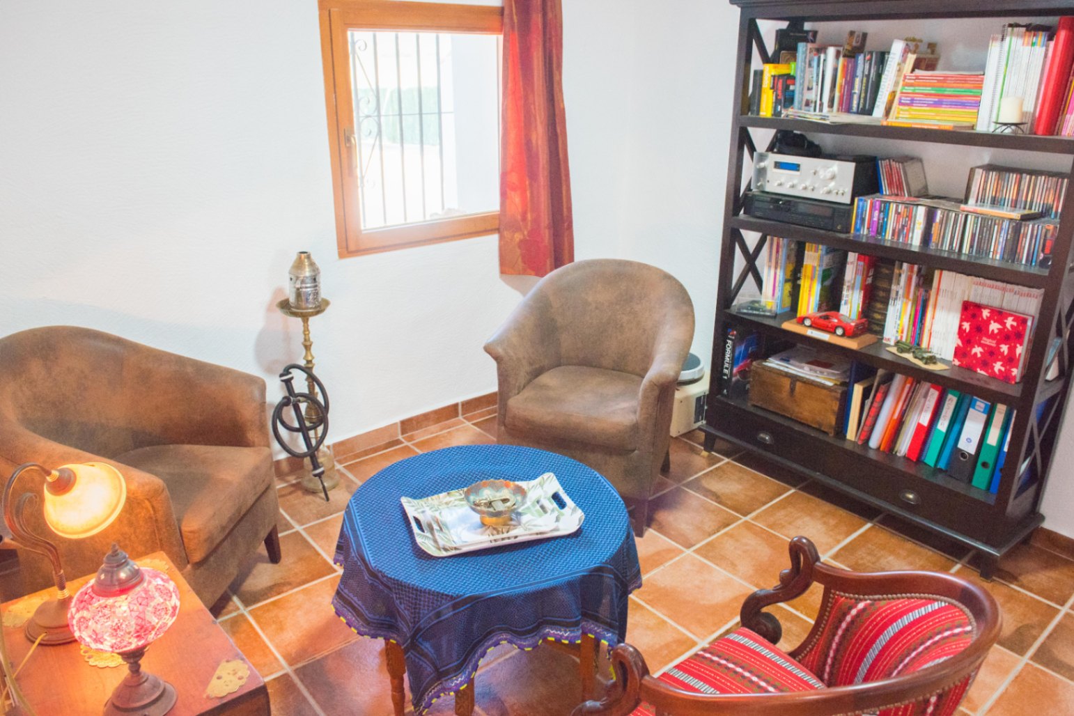 Zona de estar con suelos de baldosa de terracota, estantería de madera oscura y sillones dispuestos alrededor de una mesa circular con mantel azul.
