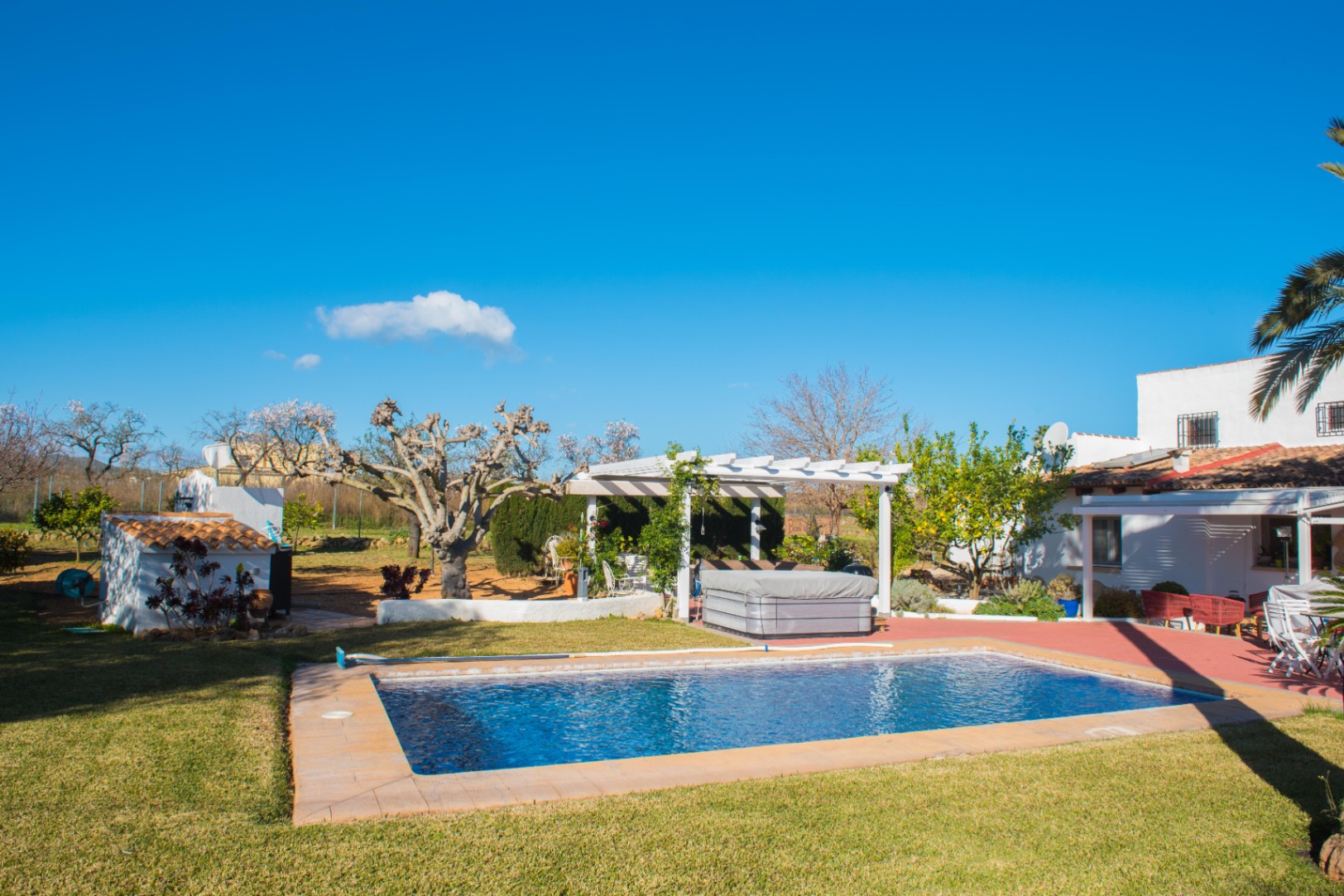 Zona exterior con piscina rectangular, césped cuidado y una pérgola blanca que resguarda un jacuzzi, integrada en un jardín de estilo mediterráneo.