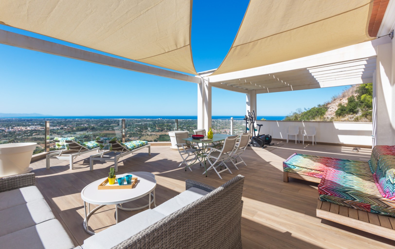 Amplia terraza con suelos de gres efecto madera, toldos de vela y vistas panorámicas al mar. Dispone de zona de comedor, sofás de exterior y tumbonas bajo una pérgola blanca.