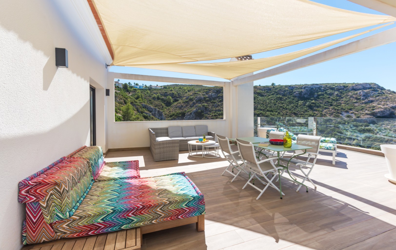 Amplia terraza con toldos vela beige, zona de comedor, sofás de mimbre y diván colorido con vistas al valle a través de barandillas de cristal.