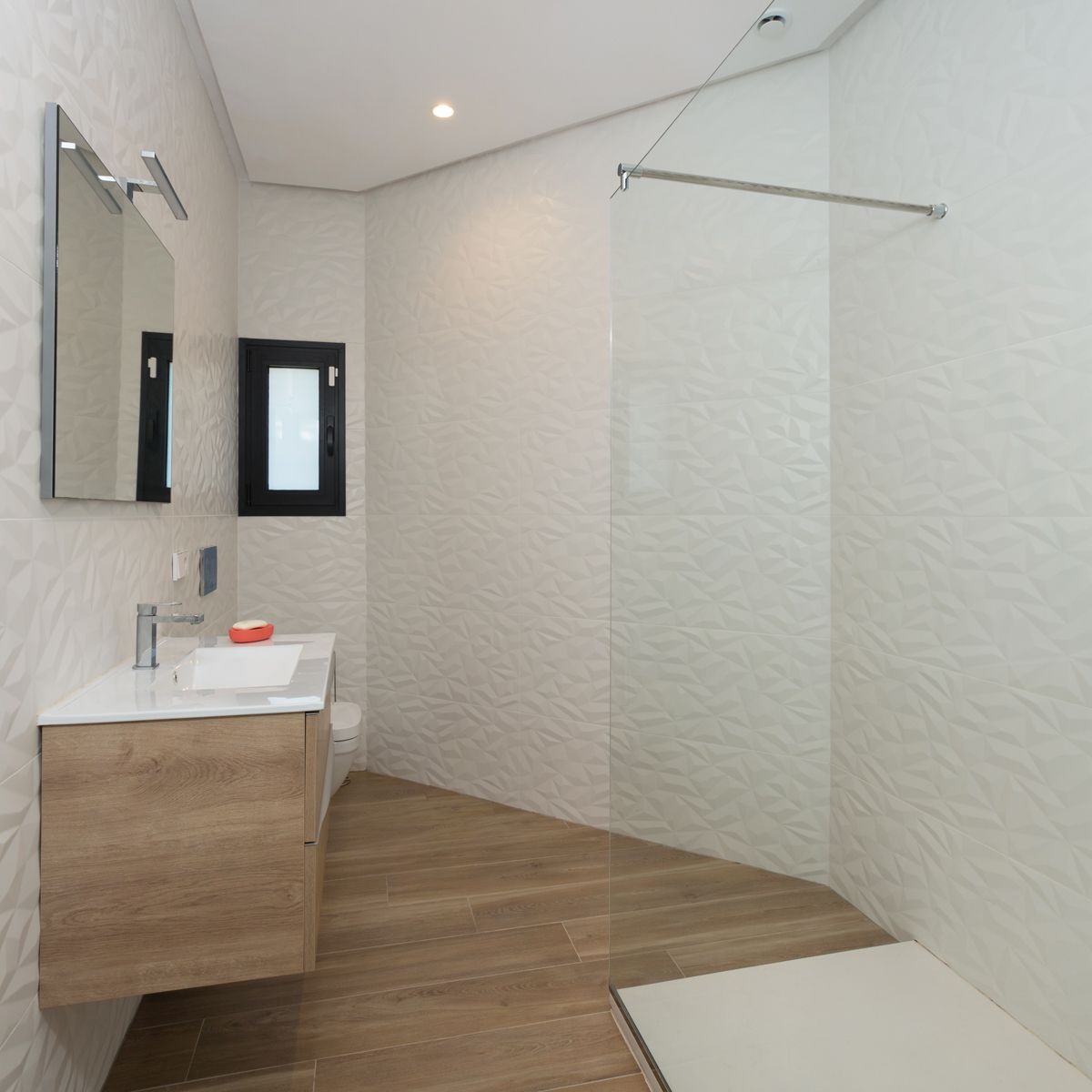 Baño contemporáneo con paredes de cerámica texturizada, mueble de lavabo suspendido en madera clara y ducha con mampara de cristal fija.