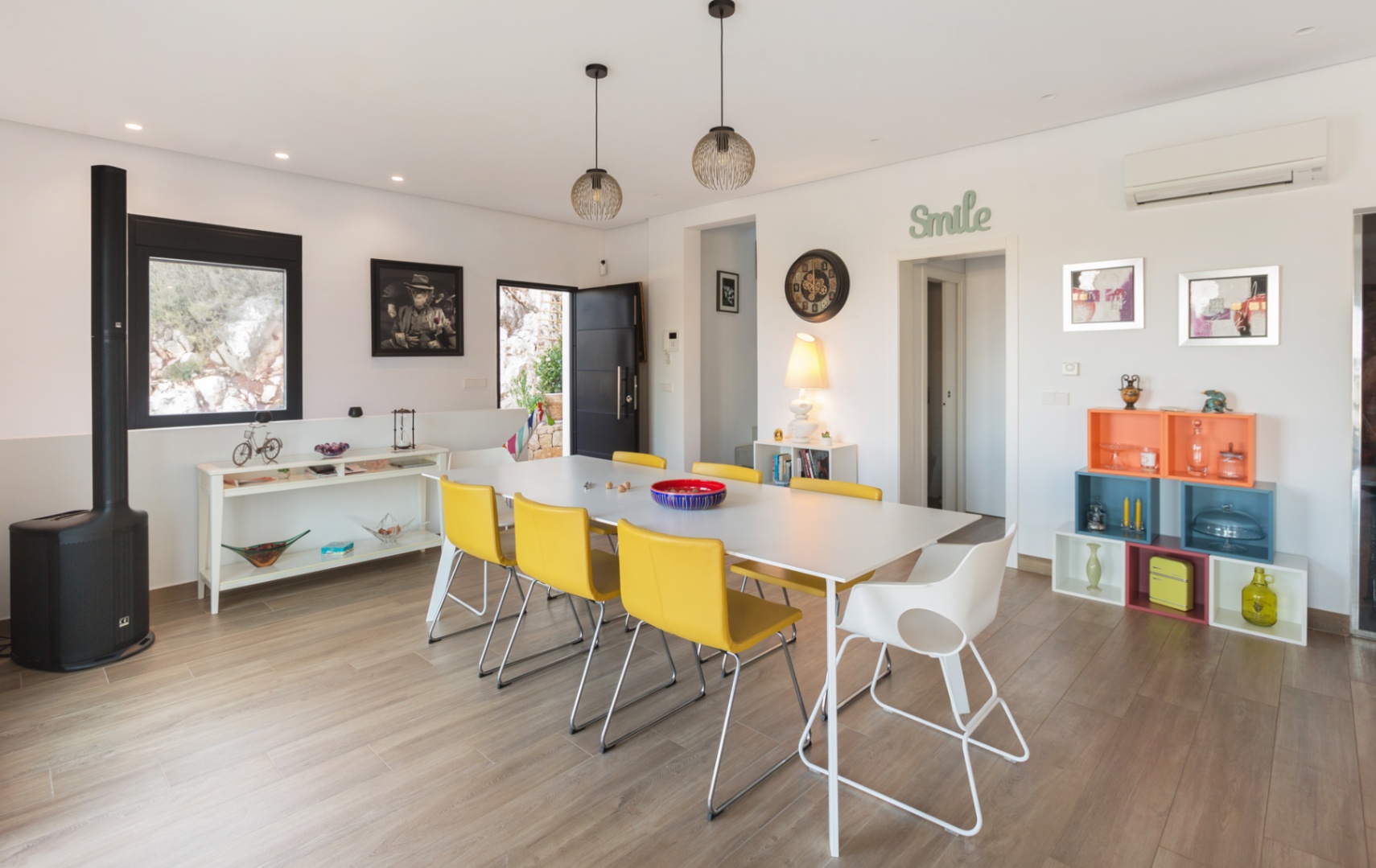 Comedor contemporáneo con mesa blanca, sillas amarillas, suelo cerámico imitación madera, estufa de leña negra y estanterías modulares de colores vibrantes.