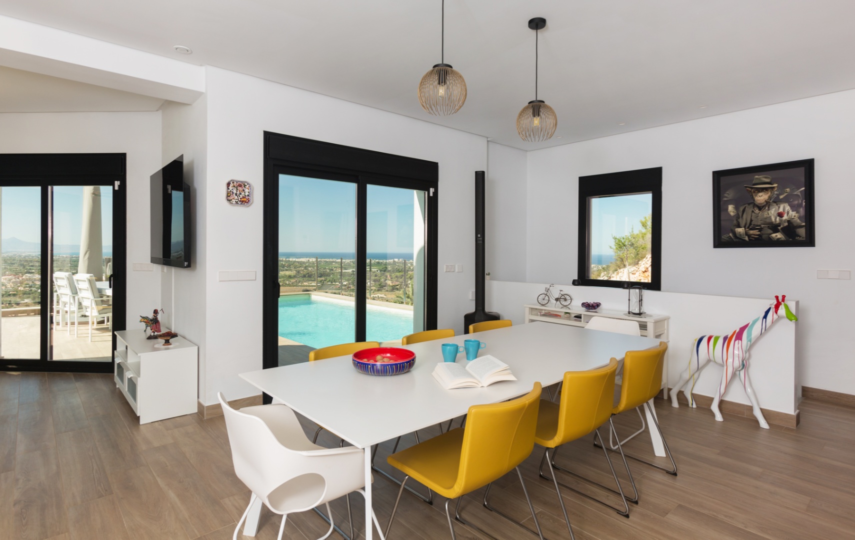Comedor moderno con mesa blanca y sillas amarillas, suelos de gres cerámico imitación madera y acceso directo a la terraza con piscina y vistas panorámicas al mar.