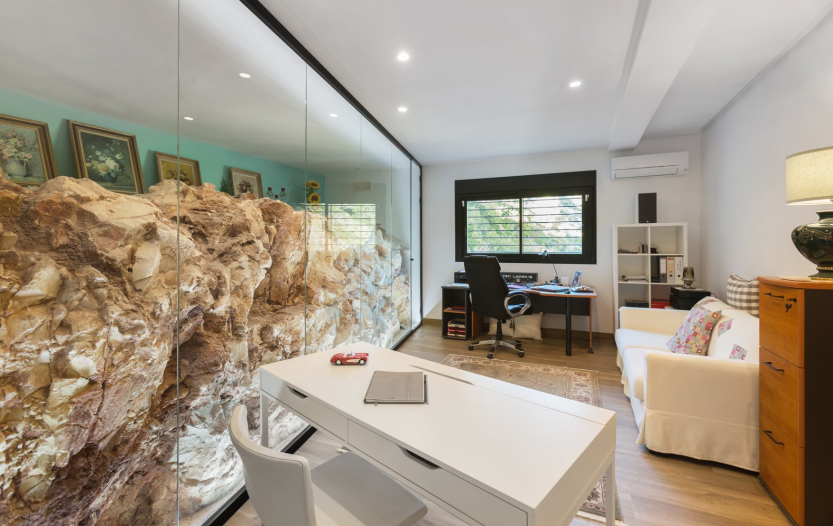 Despacho moderno con una pared de cristal que integra una formación rocosa natural. Cuenta con escritorio blanco, sofá y suelos de madera laminada.
