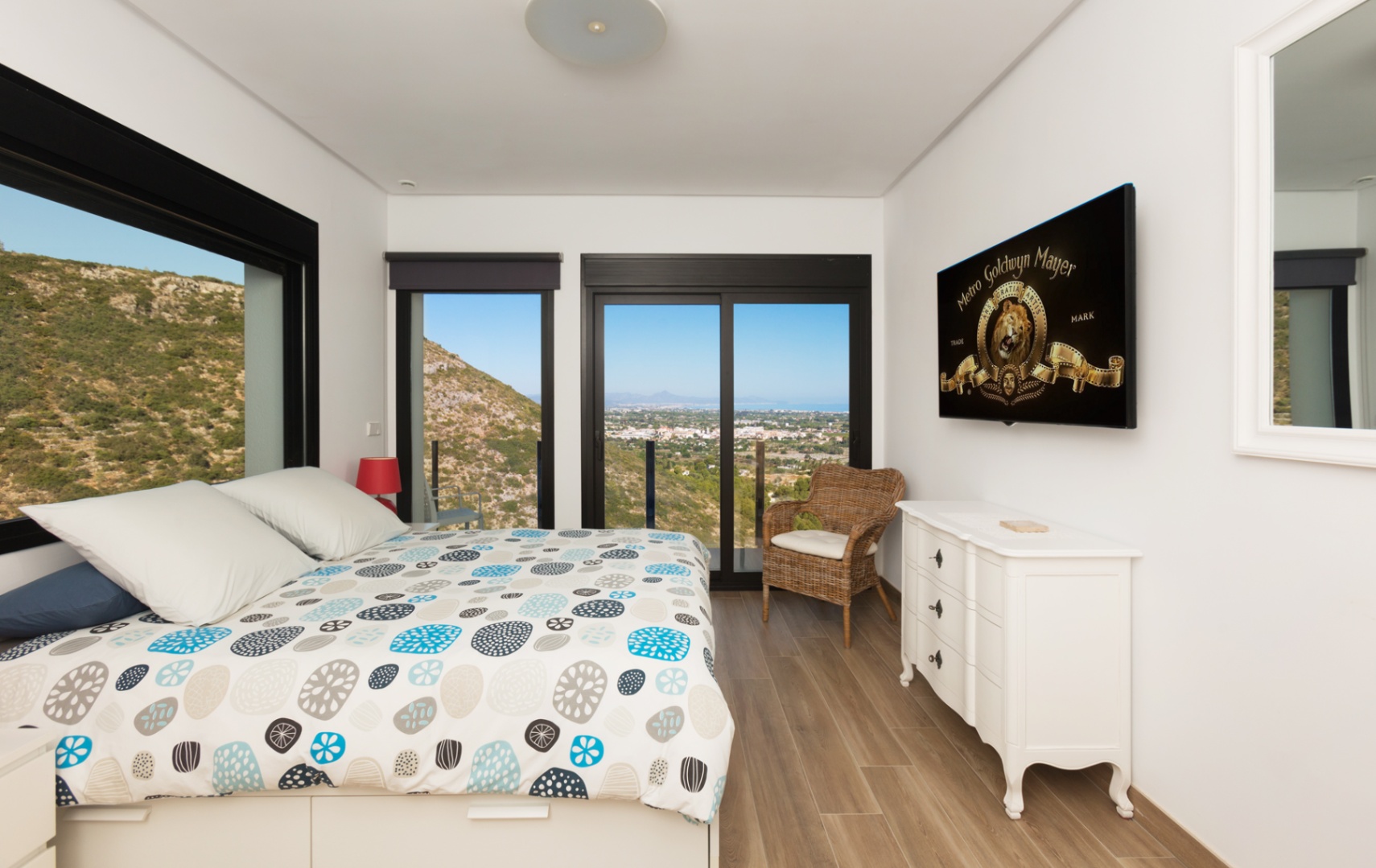 Dormitorio con vistas panorámicas a la montaña y al mar, grandes ventanales de marco negro, suelo cerámico imitación madera, cómoda blanca y sillón de mimbre.
