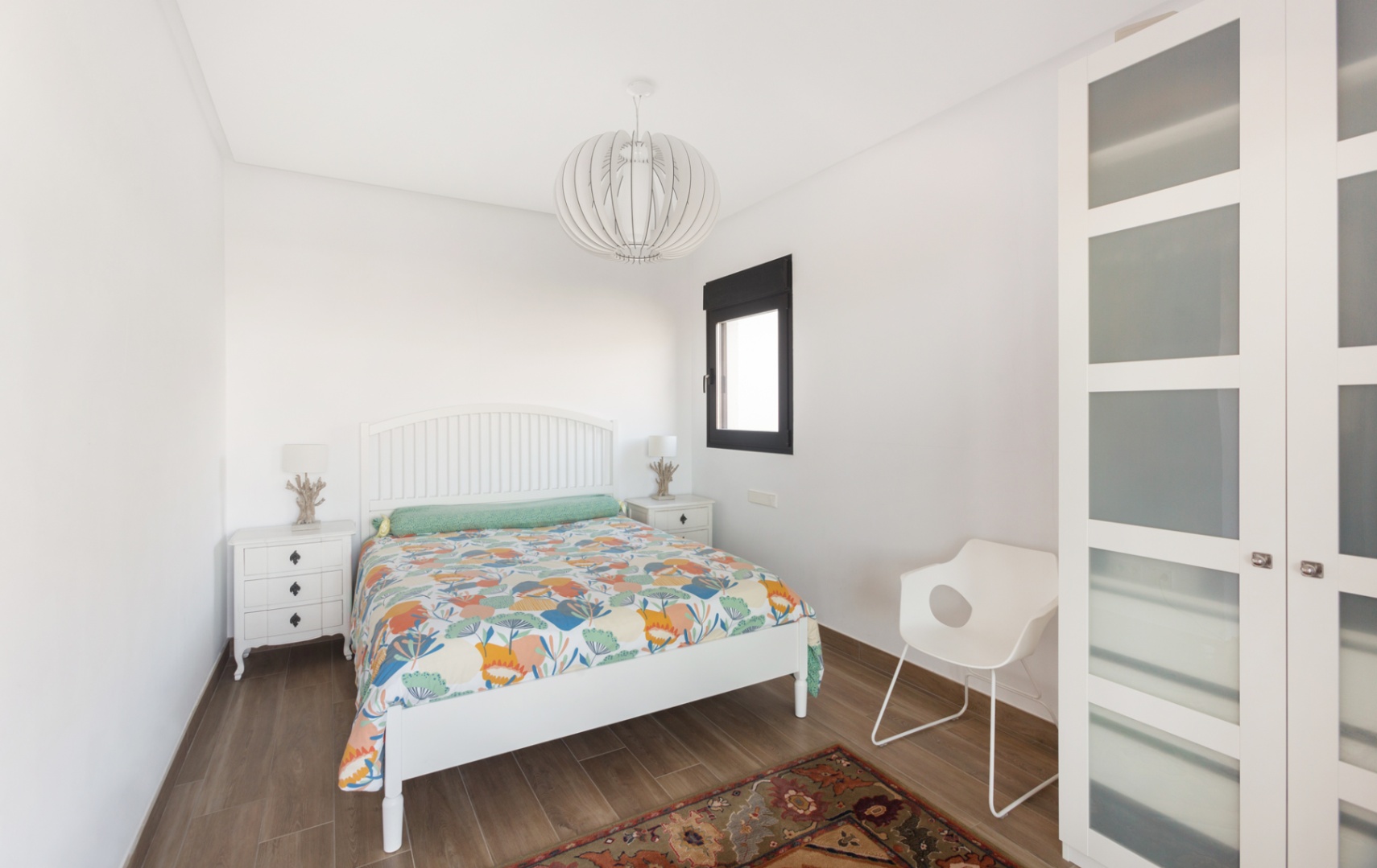 Dormitorio luminoso con cama de madera blanca, mesitas a juego y armario con paneles de cristal translúcido. Suelo de gres imitación madera y lámpara de techo de diseño.