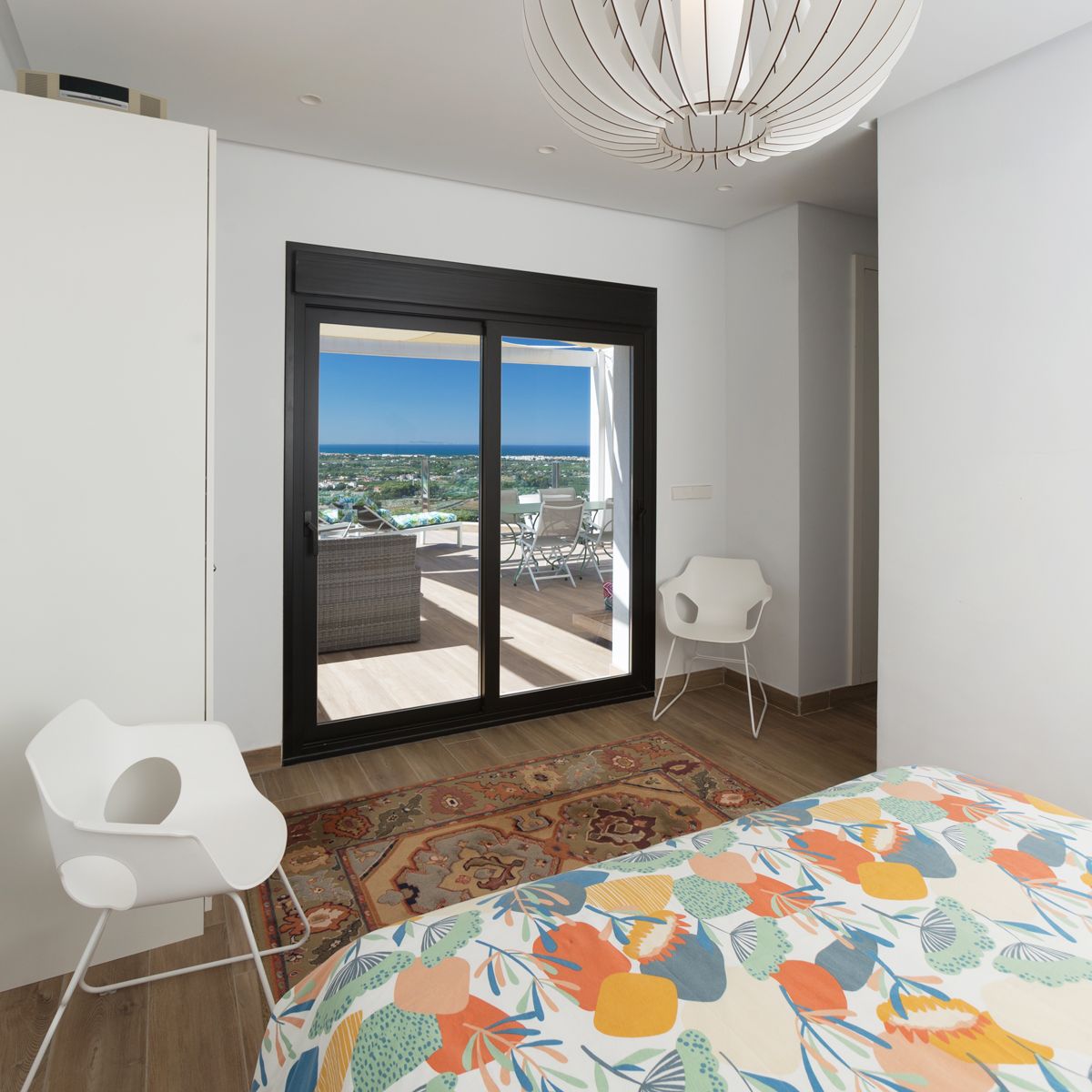 Dormitorio moderno con puerta corredera de cristal hacia una terraza amueblada con vistas al mar. Destaca la lámpara de techo escultórica y las sillas de diseño blancas.