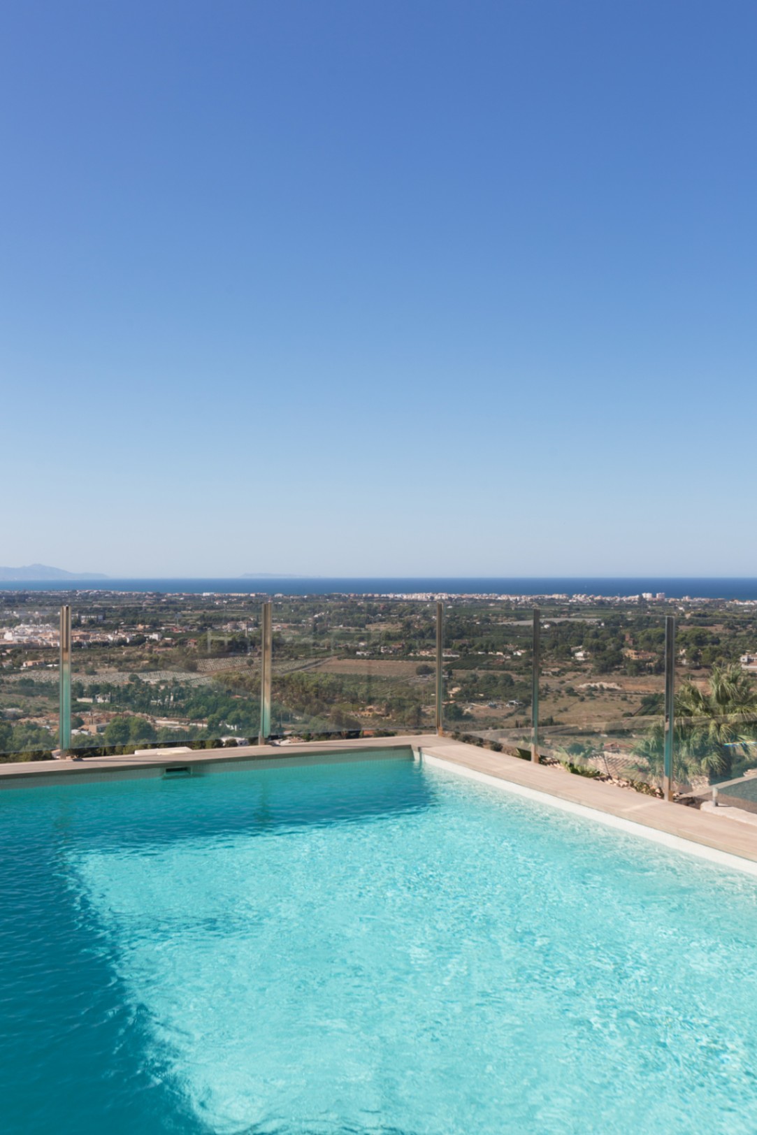 Piscina infinita elevada con barandilla de cristal, que ofrece vistas panorámicas del valle costero y el mar Mediterráneo bajo un cielo despejado.