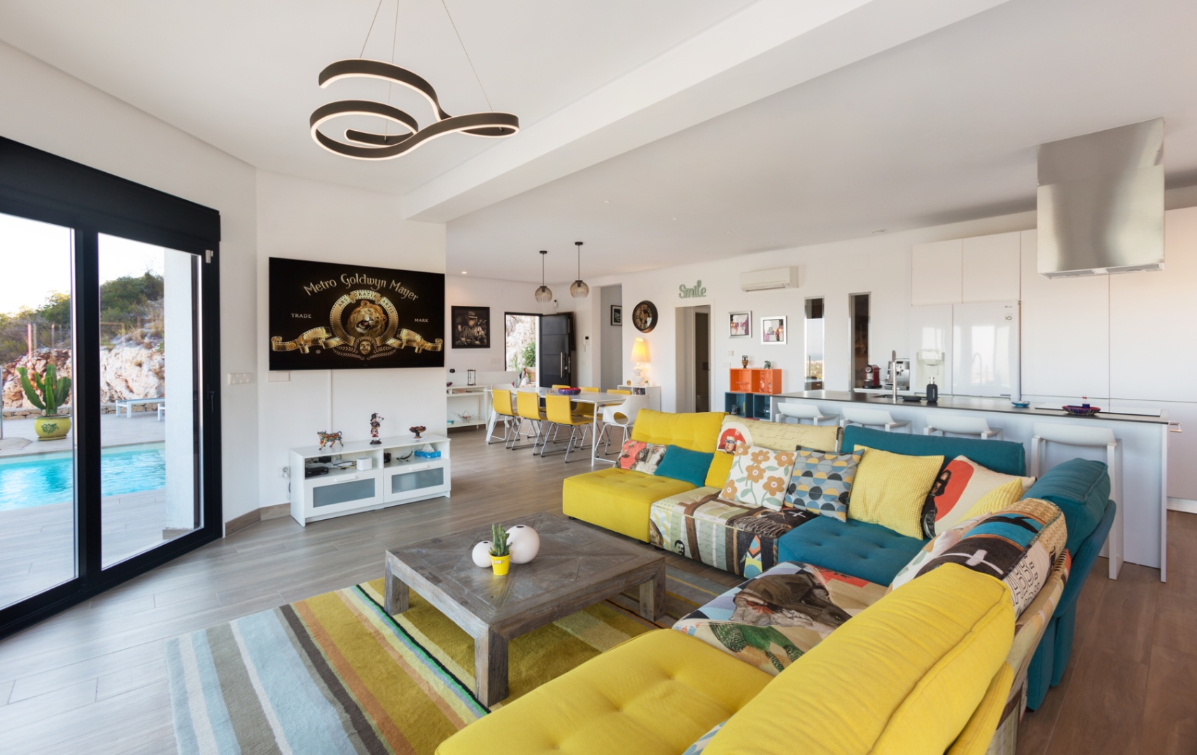Salón de concepto abierto con un sofá modular multicolor, mesa de centro de madera y acceso directo a la terraza con piscina mediante amplios ventanales.