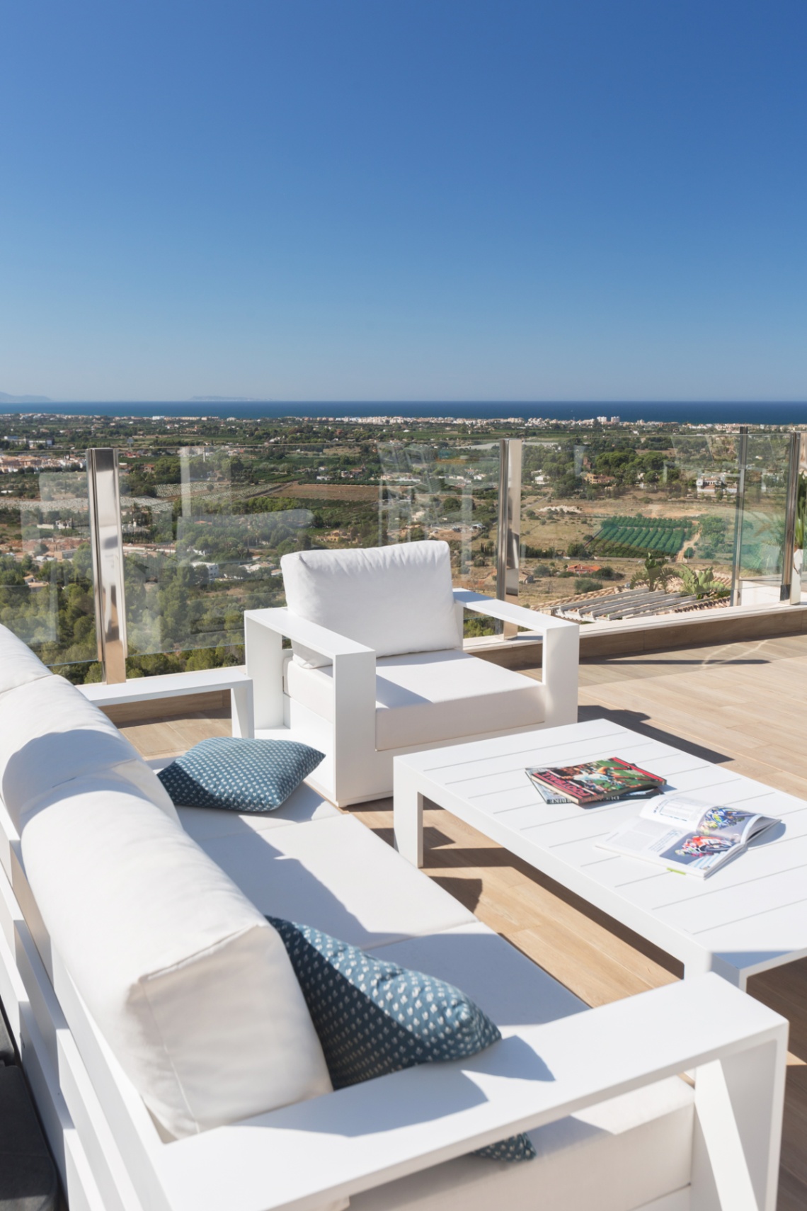 Terraza con mobiliario de exterior blanco y barandilla de cristal, con vistas panorámicas al paisaje costero y al mar Mediterráneo.