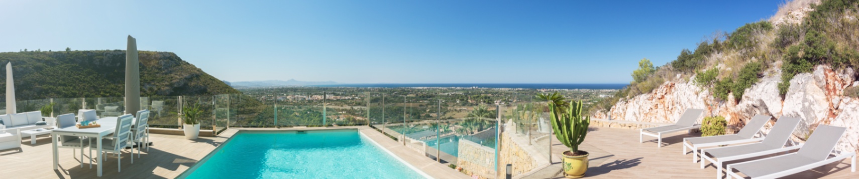 Terraza elevada con piscina, barandillas de cristal y mobiliario blanco, ofreciendo vistas panorámicas del valle y la costa mediterránea.